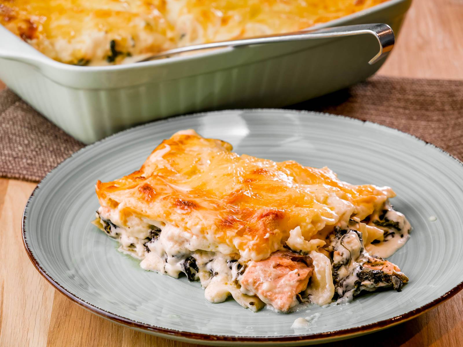 Lachs-Spinat-Lasagne Ein Stück Lachs-Spinat-Lasagne mit einer goldbraunen Käseschicht, serviert auf einem Teller. Im Hintergrund befindet sich eine Auflaufform mit der restlichen Lasagne.