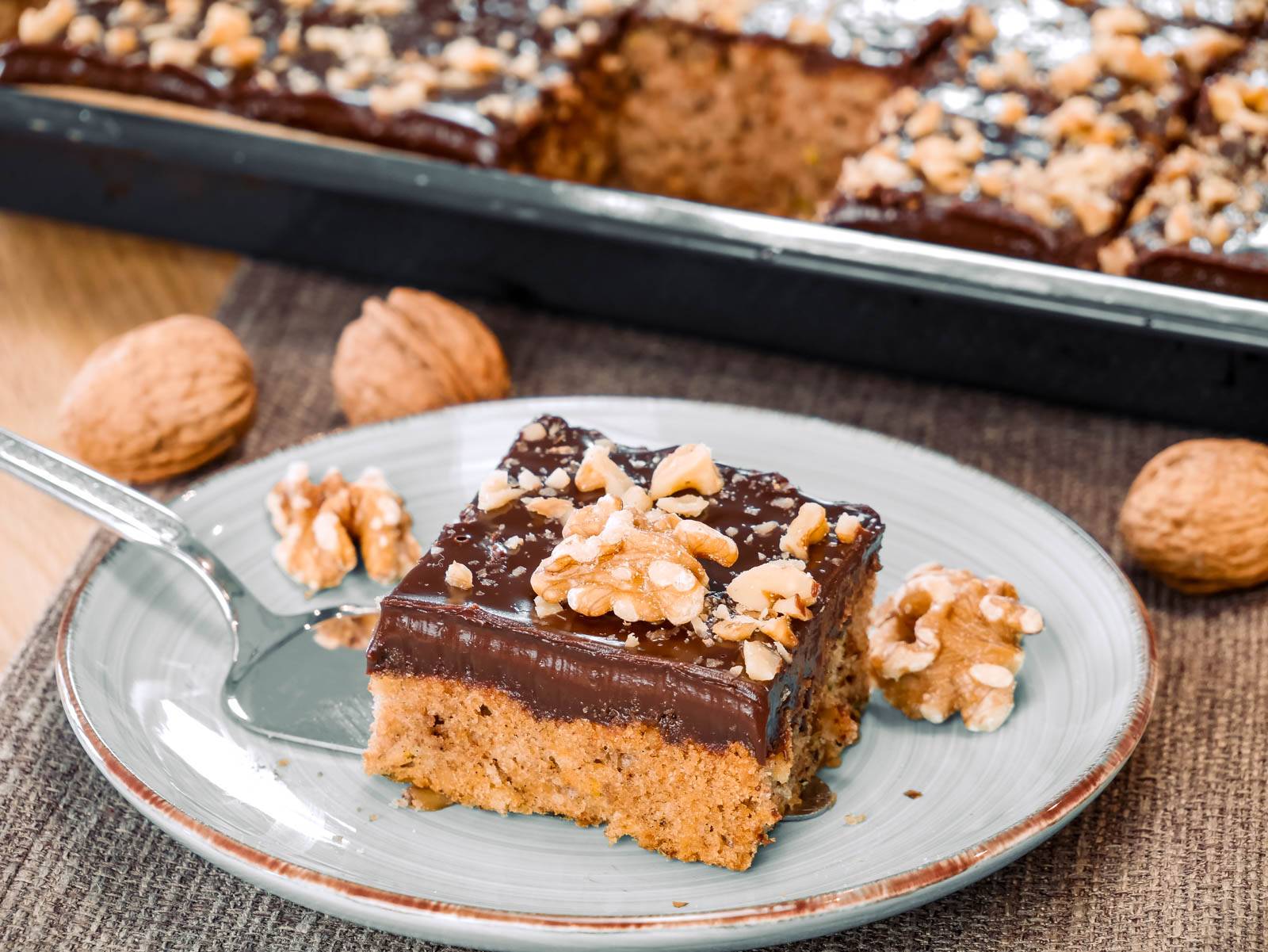 Zucchini-Nusskuchen Ein Stück Zucchini-Nusskuchen mit Schokoladenglasur und Walnüssen, serviert auf einem Teller, restlicher Kuchen im Hintergrund.