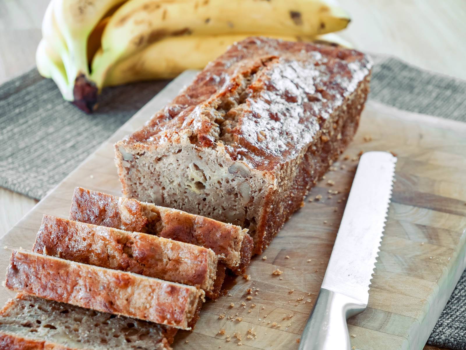 Super saftiges Bananabread Ein frisch gebackenes Bananenbrot, in Scheiben geschnitten und auf einem Holzbrett serviert. Im Hintergrund liegen reife Bananen.