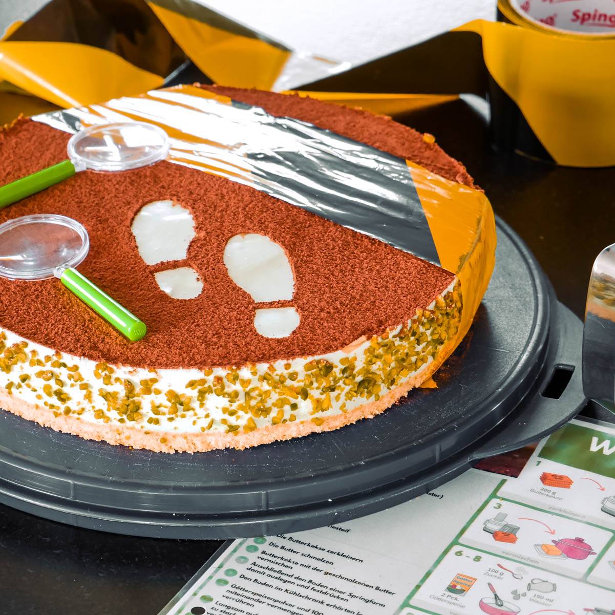 Waldmeister-Philadelphia-Torte Philadelphia-Torte mit Kakaoschicht und Fußabdruck-Muster, dekoriert mit zwei grünen Lupen und gelb-schwarzem Band, Bildrezept im Hintergrund.