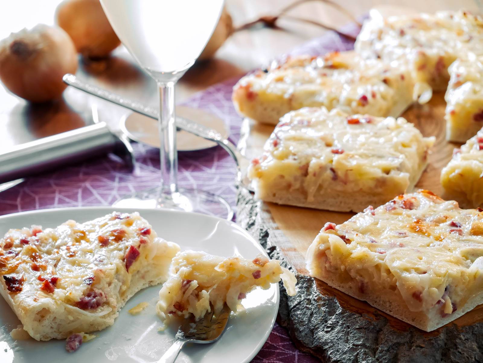 Zwiebelkuchen vom Blech mit Käse – Einfach und Lecker Ein Stück Zwiebelkuchen, belegt mit geschmolzenem Käse und Speck, serviert auf einem Teller mit einer Gabel.