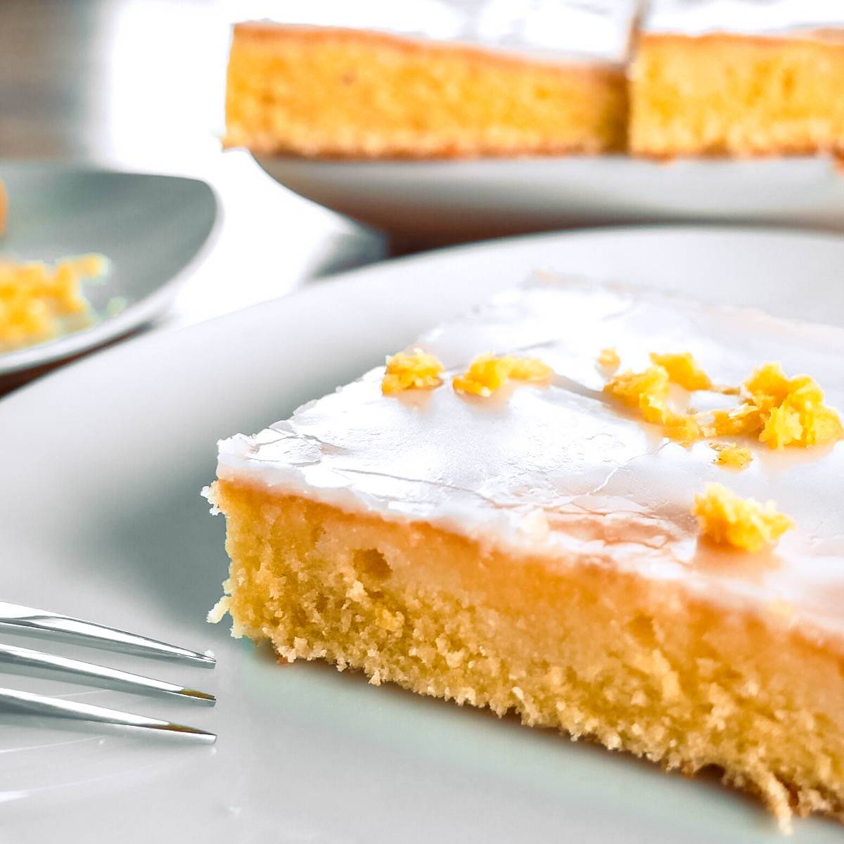 Zitronenkuchen Ein Stück Zitronenkuchen mit Zuckerguss auf einem Teller, der restliche Kuchen im Hintergrund.