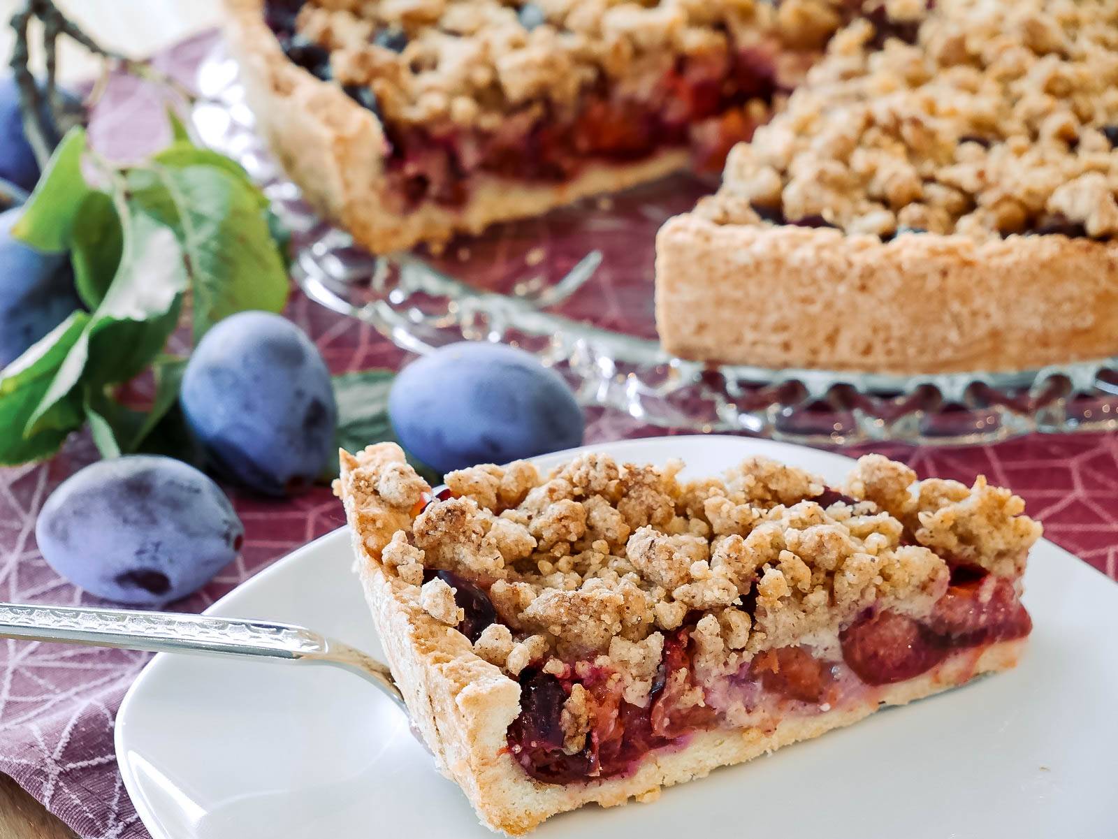 Zwetschgenkuchen mit Mürbteig und Streusel Ein Stück Zwetschgenkuchen mit Streuseln auf einem Teller. Im Hintergrund ist der restliche Kuchen auf einer Glasplatte sowie frische Zwetschgen zu sehen.
