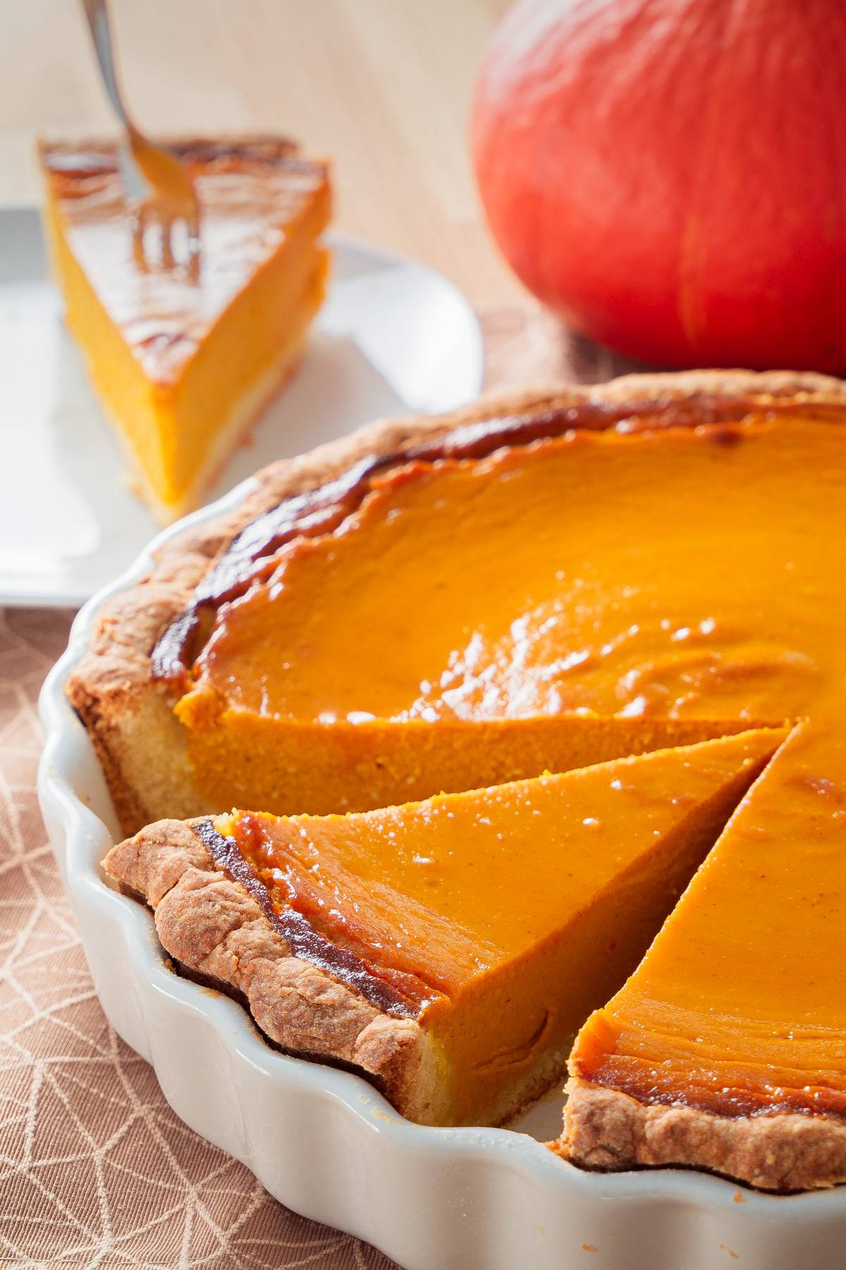 Ein Stück cremiger Pumpkin Pie auf einem Teller, mit einem Hokkaido-Kürbis und der restlichen Tarte im Vordergrund.