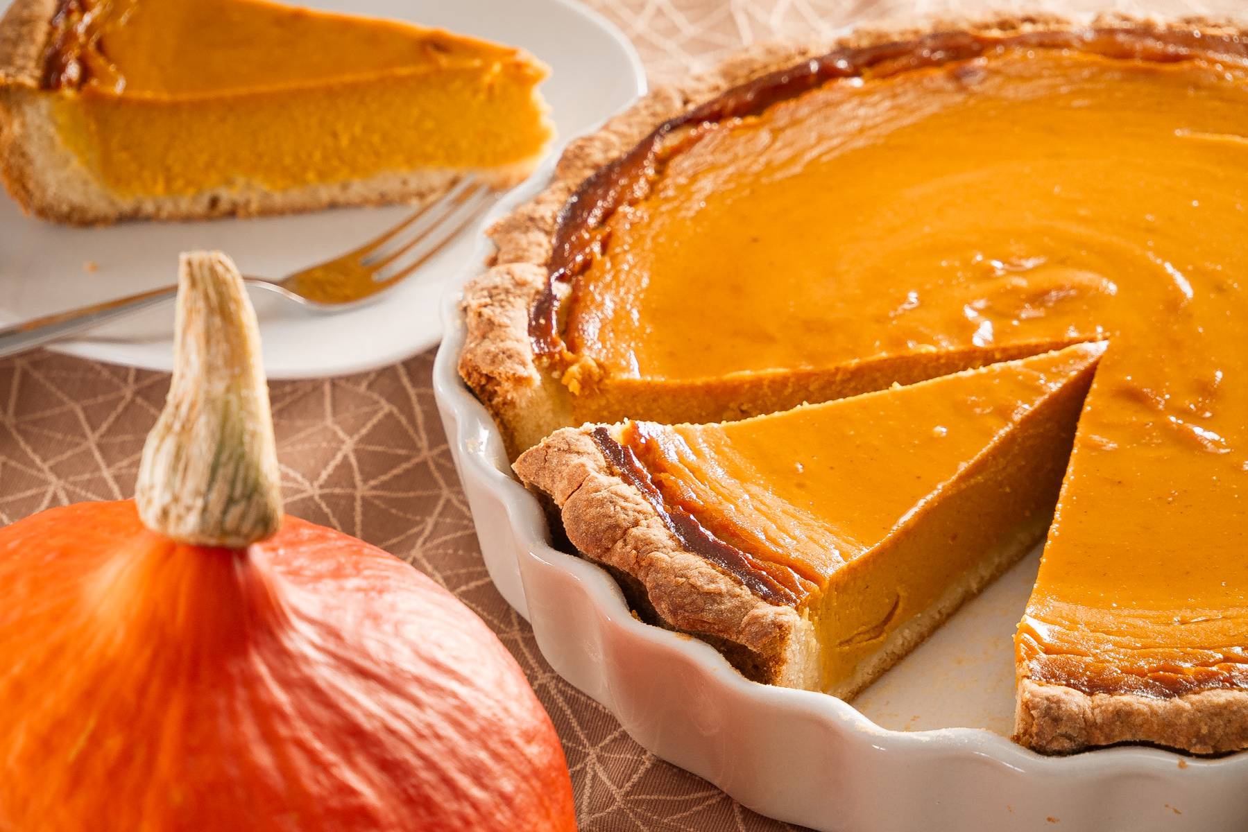 Leckeres Pumpkin Pie Rezept – Der amerikanische Klassiker Ein Stück Pumpkin Pie auf einem Teller, mit einem Hokkaido-Kürbis im Vordergrund und der restlichen Tarte im Hintergrund.