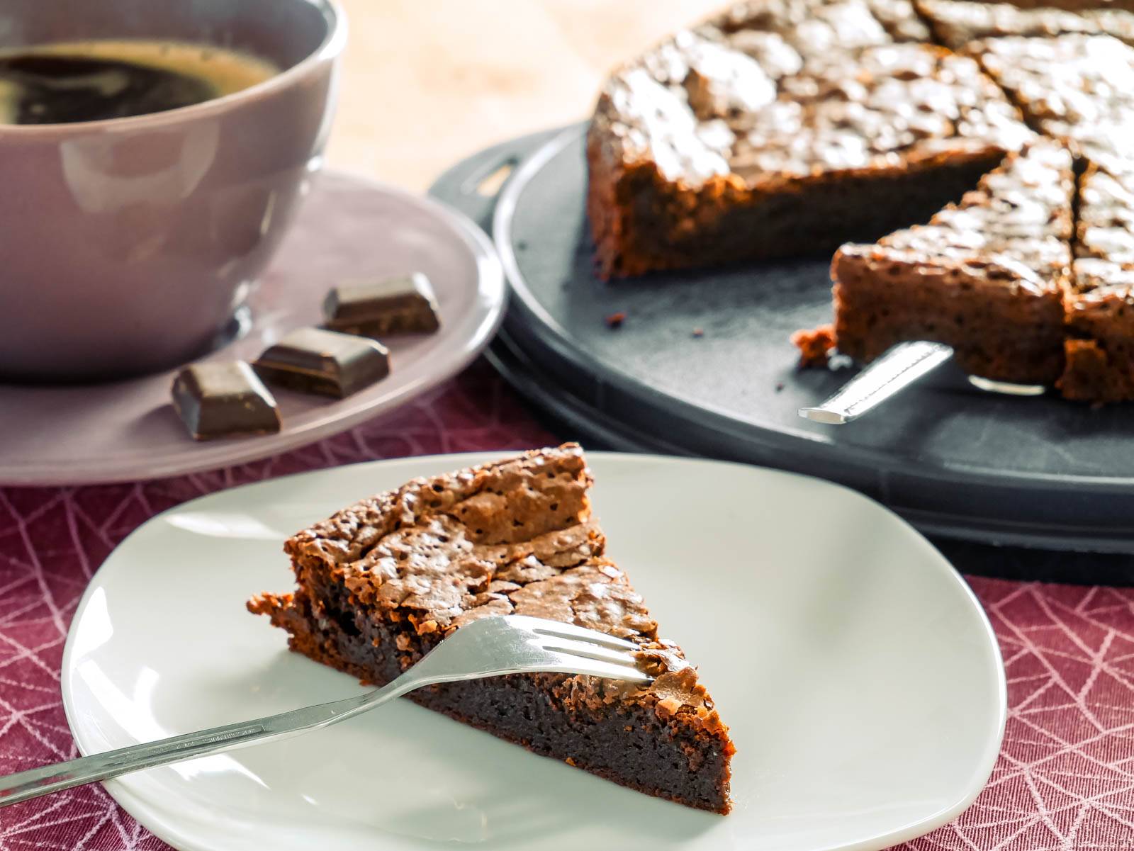Tarte au Chocolat Ein Stück Tarte au Chocolat auf einem Teller, daneben eine Tasse Kaffee und einige Schokoladenstücke. Im Hintergrund steht der restliche Kuchen auf einer Kuchenplatte.