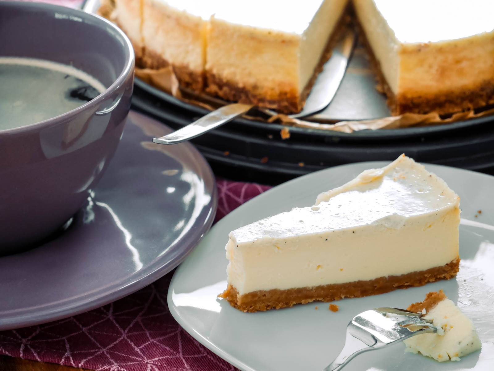New York Cheese Cake Ein Stück New York Cheesecake auf einem Teller mit einer Gabel daneben. Im Hintergrund steht der restliche Kuchen auf einer Kuchenplatte.