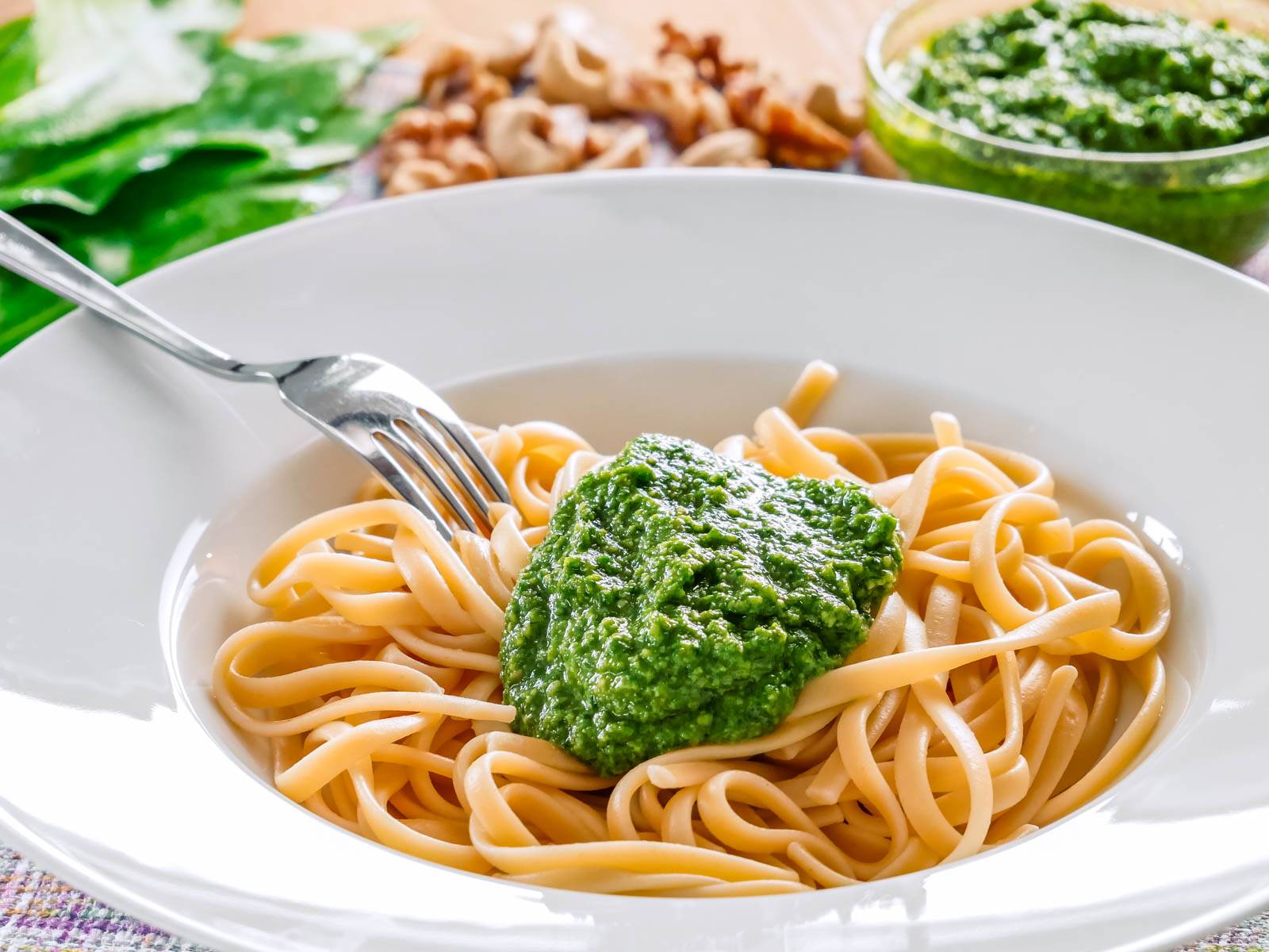 Bärlauch – Der Knoblauch des Waldes Ein Teller mit Spaghetti und einem Klecks grünem Bärlauch Pesto, garniert mit einer Gabel, die teilweise unter der Pasta verborgen ist, auf einem Tisch mit unscharfem Hintergrund.