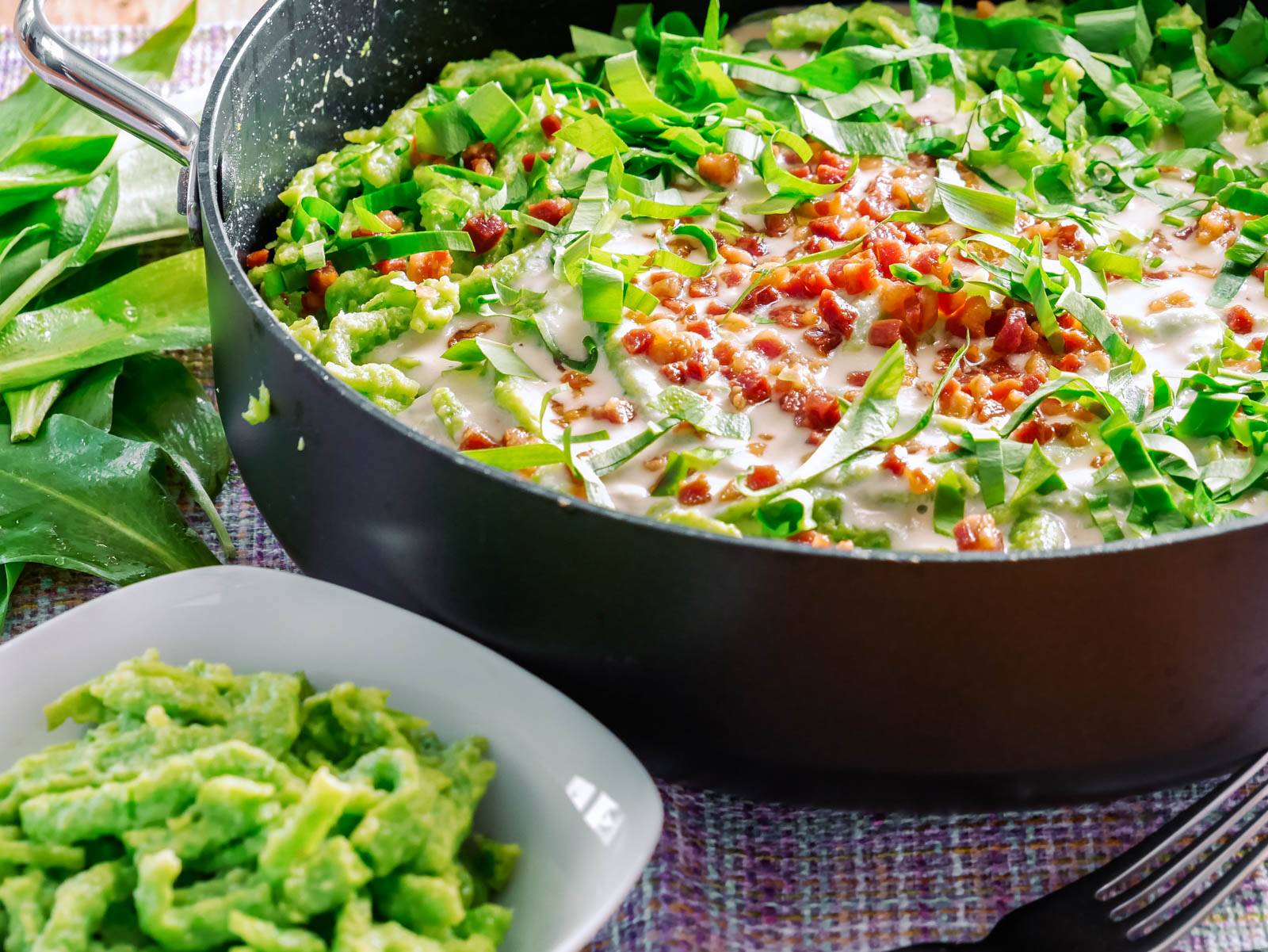 Bärlauch – Der Knoblauch des Waldes Bärlauchspätzle in einer Pfanne mit cremiger Gorgonzolasauce, bestreut mit Speck, serviert mit einer Schale weiterer Spätzle.