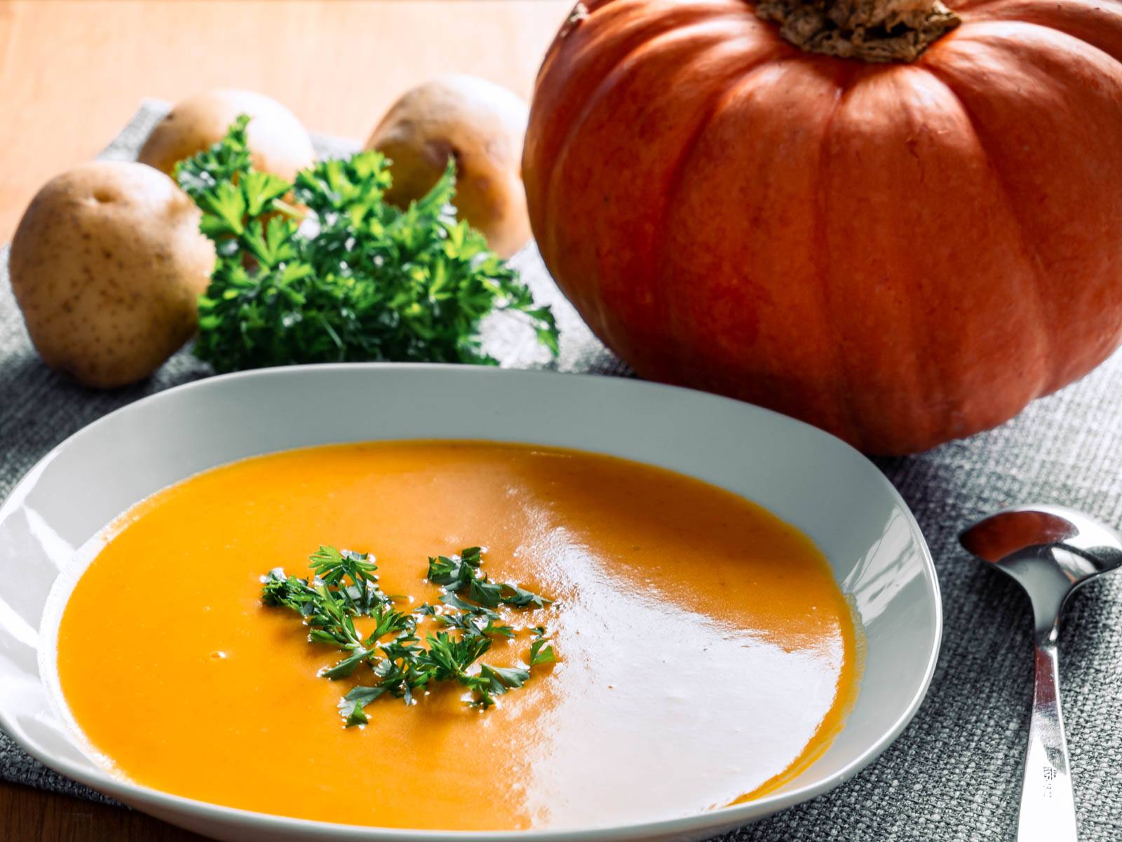 Ein Teller mit cremiger Kürbissuppe, garniert mit frischer Petersilie. Im Hintergrund liegen Kartoffeln, ein großer Kürbis und ein Bund Petersilie auf einer grauen Tischdecke. Ein Löffel liegt neben dem Teller.