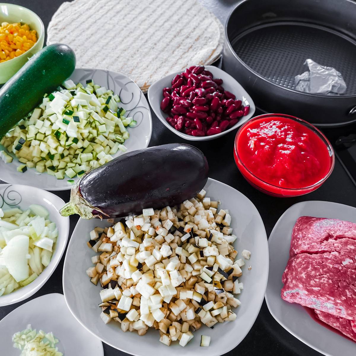 Mexikanische Tortilla Lasagne – Der perfekte Wrap Auflauf für alle Tex-Mex-Fans! Zutaten für Mexikanische Tortilla Lasagne, einschließlich Hackfleisch, Bohnen, Mais, Zucchini, Aubergine und Tomatensauce, schön arrangiert auf einem Tisch.