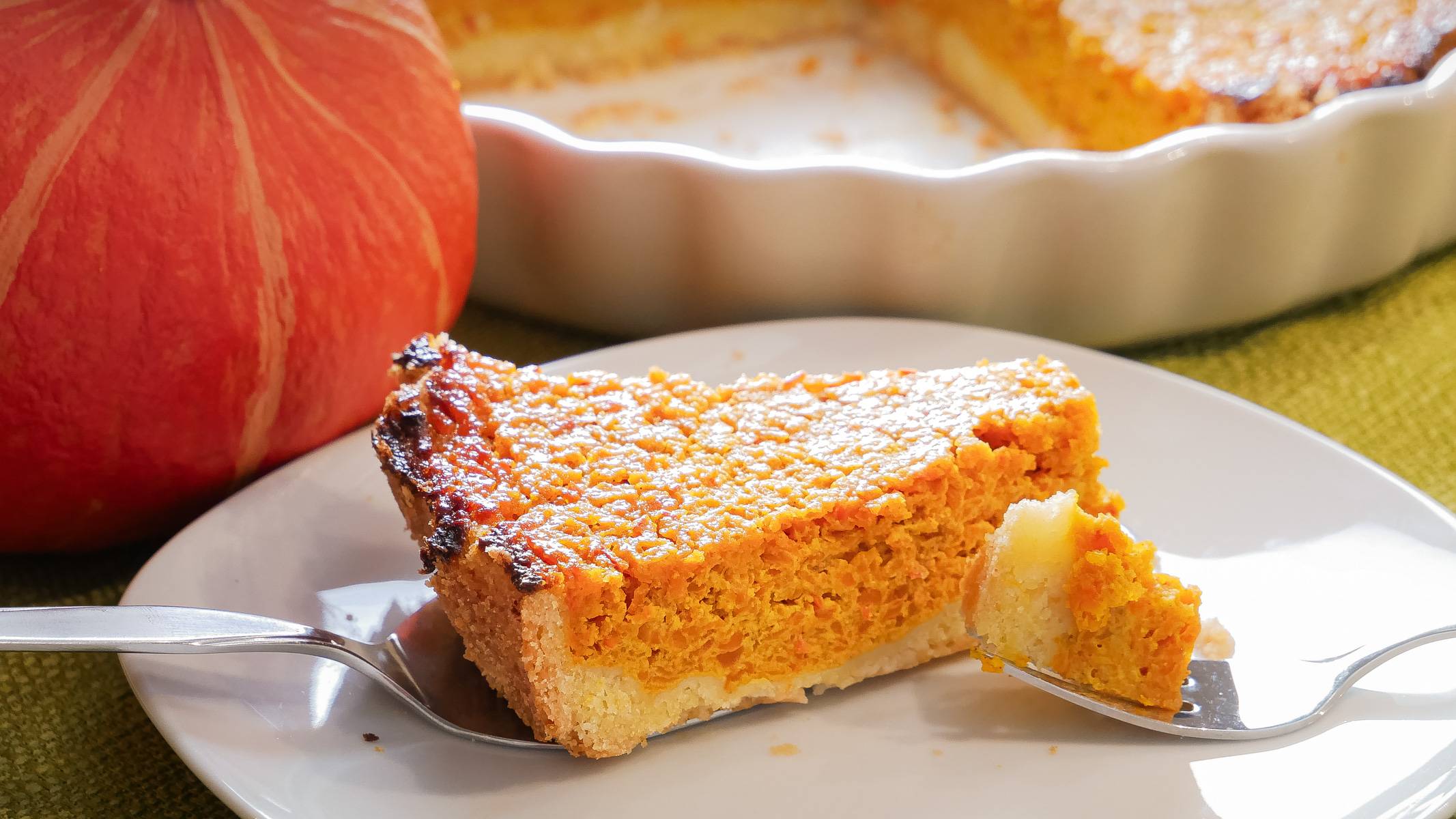 Süße Kürbistarte - herbstliches Rezept für Kaffee und Tee Ein Stück süße Kürbistarte auf einem Teller mit einer Gabel, im Hintergrund die restliche Tarte in einer weißen Backform und ein Hokkaido-Kürbis als Dekoration.