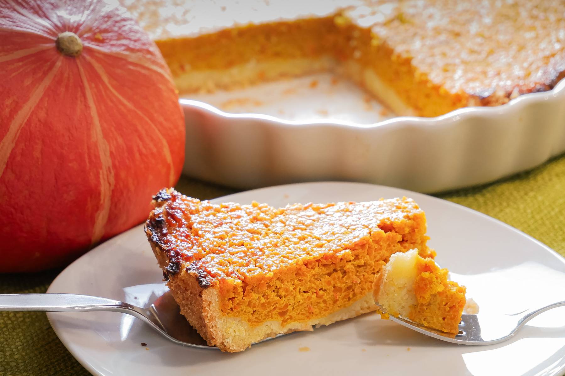 Süße Kürbistarte - herbstliches Rezept für Kaffee und Tee Ein Stück süße Kürbistarte auf einem Teller mit einer Gabel, im Hintergrund die restliche Tarte in einer weißen Backform und ein Hokkaido-Kürbis als Dekoration.